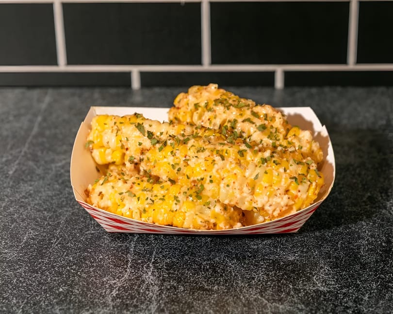 Elote-style corn ribs, coated in creamy sauce and sprinkled with herbs, in a striped paper tray.