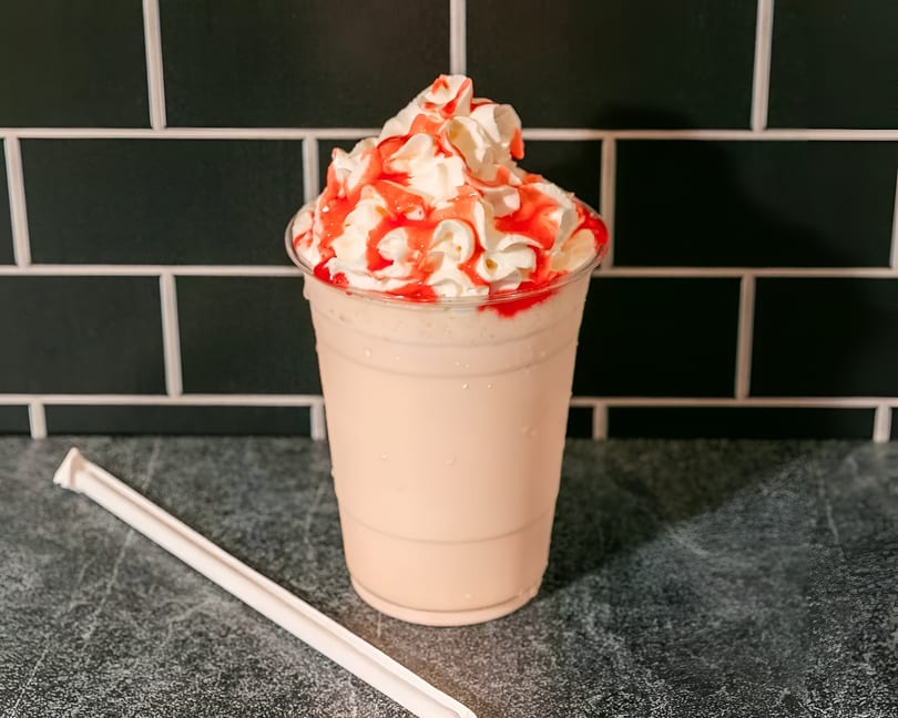 A strawberry milkshake with whipped cream and red syrup drizzle, next to a white straw.