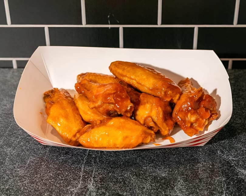 A white paper tray with red stripes holds a pile of saucy, orange-glazed chicken wings.