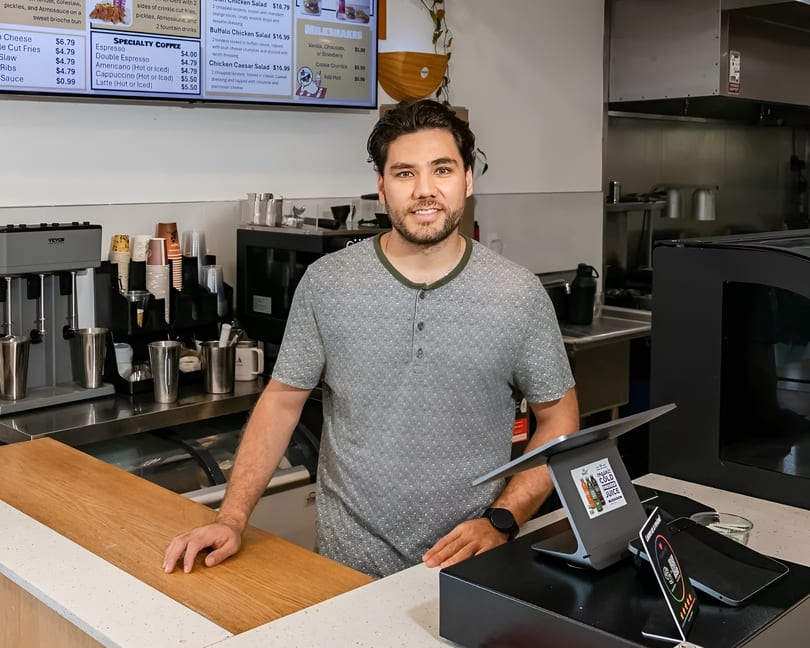 A smiling male barista stands behind a counter with coffee machines and a digital menu board.