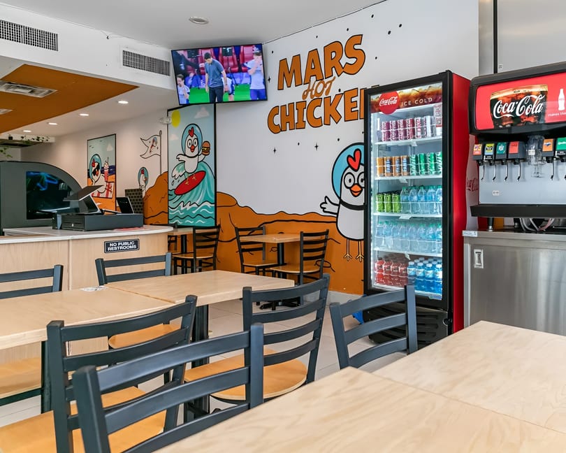 Interior of Mars Hot Chicken restaurant with tables, chairs, colorful wall murals, and a drink station.
