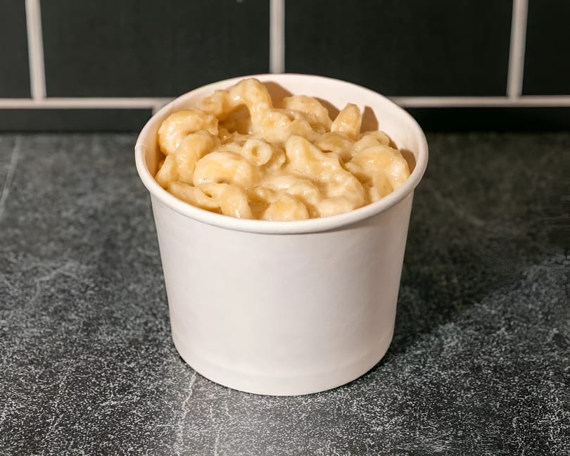 Creamy mac and cheese in a white to-go cup on a dark speckled counter with a black tiled wall.